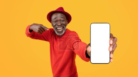 Téléchargez les photos : Heureux homme noir senior pointant vers un écran de smartphone vierge avec un sourire rayonnant, portant un pull rouge et un chapeau, parfait pour la promotion de l'application, isolé sur un fond jaune - en image libre de droit