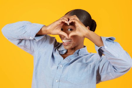 Téléchargez les photos : Drôles afro-américaine jeune dame regardant la caméra à travers coeur en forme de doigts geste, sur fond jaune studio, flirt et publicité Sortir ensemble Service. J'aime ça Concept - en image libre de droit