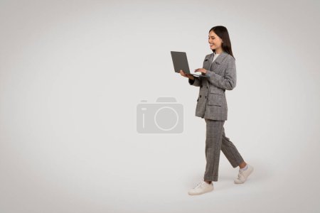Téléchargez les photos : Jeune femme professionnelle active marchant et utilisant un ordinateur portable, vêtue d'un costume de pied-de-poule avec des baskets décontractées, sur fond gris minimaliste, espace libre - en image libre de droit