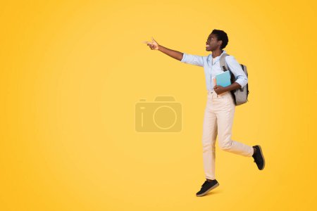 Téléchargez les photos : Heureuse jeune étudiante afro-américaine décontractée avec sac à dos, les promenades de livres apprécient le temps libre, pointent du doigt l'espace de copie, isolée sur fond de studio jaune. Etude et éducation, annonce, offre - en image libre de droit