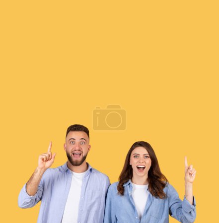Téléchargez les photos : Joyeusement surpris homme et femme pointant vers le haut vers l'espace libre, partageant un moment eureka enthousiaste, à la fois dans des chemises bleues décontractées sur fond jaune joyeux - en image libre de droit