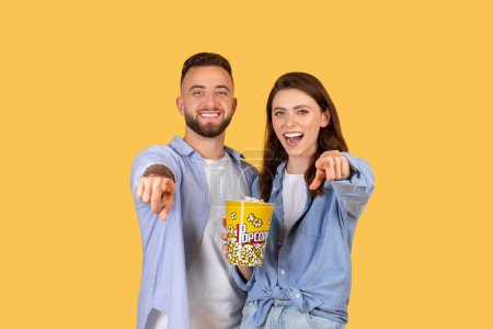 Téléchargez les photos : Joyeux jeune homme et femme pointant vers le spectateur, partageant seau de maïs soufflé, exprimant l'excitation et le plaisir sur fond jaune - en image libre de droit
