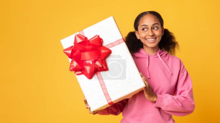 Téléchargez les photos : Cadeau d'anniversaire. Jeune fille noire curieuse secouant grande boîte, vérifier cadeau tout en posant sur fond jaune studio, panorama, espace libre. Offres vacances et célébration - en image libre de droit