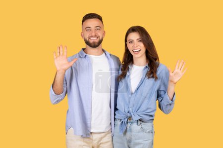 Photo for Cheerful young couple in casual blue shirts waving their hands in friendly greeting, showing openness and welcoming attitude on yellow backdrop - Royalty Free Image