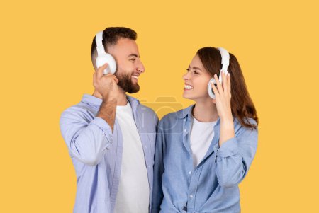 Téléchargez les photos : Un homme et une femme joyeux profitent d'une expérience musicale partagée, chacun portant un casque blanc sur l'oreille et se souriant sur fond jaune - en image libre de droit