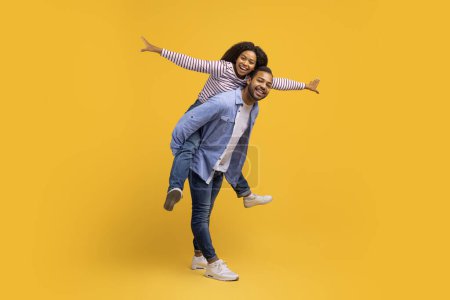 Photo for Loving young black couple enjoying time together, having fun, cheerful african american woman piggybacking her boyfriend, imitating airplane, posing over yellow studio background, copy space - Royalty Free Image
