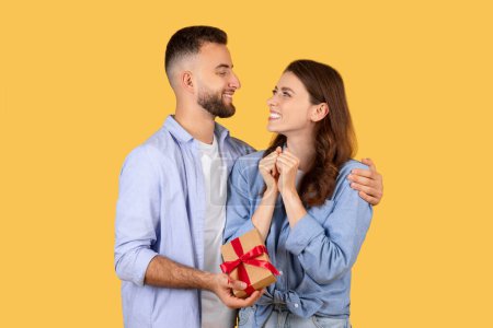 Photo for Happy couple sharing special moment, as smiling man surprises his partner with wrapped gift, eliciting an excited and grateful reaction from her - Royalty Free Image