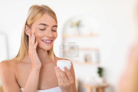 Téléchargez les photos : Jeune femme blonde européenne dorlote sa peau avec des cosmétiques, applique crème faciale embrassant la routine de soins de la peau domestique, exsudant bien-être et mode de vie heureux dans une salle de bain confortable à l'intérieur. Espace libre - en image libre de droit