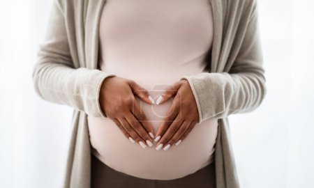 Téléchargez les photos : Coupé de femme enceinte noire tenant la main sur son gros ventre, signe en forme de cœur, isolé sur fond blanc, gros plan. Grossesse, maternité, maternité - en image libre de droit