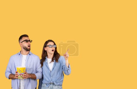 Téléchargez les photos : Incroyable homme et femme portant des lunettes 3D, regardant et pointant vers le haut avec sens de l'émerveillement, tenant seau de maïs soufflé sur fond jaune - en image libre de droit