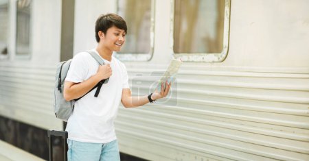Foto de Joven asiático llevando mochila turista chequeando mapa de papel, de pie en tren en la estación, mirando el punto de destino, panorama con espacio de copia. Viajar, turismo, vacaciones - Imagen libre de derechos