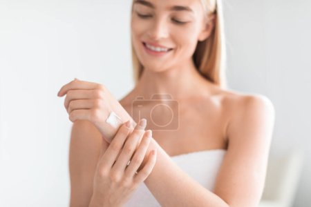Photo for Attractive blonde young lady in towel applying lotion to her arms, focusing on moisturizing dry skin in her home bathroom. Moisturizer advertisement. Cropped shot, selective focus on hands - Royalty Free Image