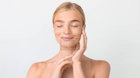 Photo for Beauty portrait of young blonde lady enjoying skincare routine, hands gently touching her face against white backdrop in studio. Headshot of woman closing eyes posing elegantly. Panorama - Royalty Free Image