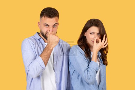 Photo for Man and woman in casual shirts make faces of disgust and pinch their noses, showing strong reaction to bad smell on plain yellow backdrop - Royalty Free Image
