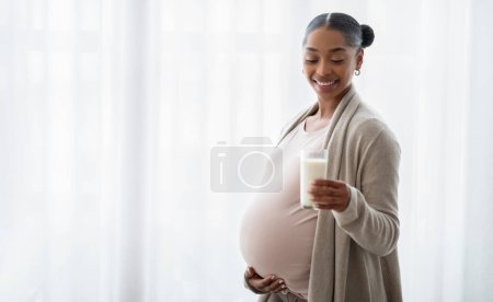 Photo for Lactation and breastfeeding concept. Pregnant black woman standing next to window at home, hugging her belly and offering glass with fresh milk on camera, panorama with copy space - Royalty Free Image