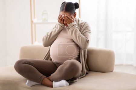 Foto de Cambios hormonales del embarazo. Mujer afroamericana embarazada joven sintiéndose triste, sentada sola en el sofá y llorando en casa, cubra su cara con palmas, espacio libre - Imagen libre de derechos