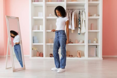 Photo for Sad curly-haired latin woman critically assessing the fit of new blue jeans while wearing snug white t-shirt in her pink-toned minimalist room - Royalty Free Image