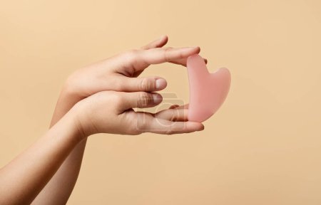 Foto de Las manos muestran elegantemente la herramienta rosa facial de cuarzo gua sha en forma de corazón contra un fondo beige suave, que simboliza la belleza y las rutinas de cuidado de la piel en casa, recortadas, de cerca - Imagen libre de derechos