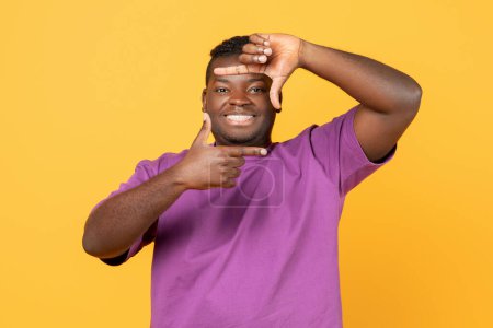 Téléchargez les photos : Joyeux homme afro-américain en t-shirt violet faisant cadre photo avec les doigts, capturant des moments et souriant à la caméra, debout sur fond de studio jaune. Concept de photographie - en image libre de droit