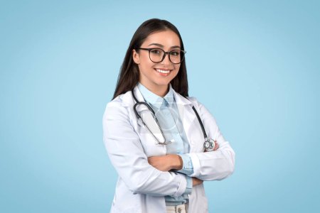 Foto de Retrato de una joven profesional de medicina general en gafas posando con los brazos cruzados sobre fondo azul del estudio, sonriendo a la cámara - Imagen libre de derechos