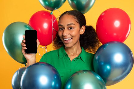 Téléchargez les photos : Heureuse adolescente noire dans des poses décontractées avec téléphone portable parmi les ballons flottants, offrant un écran vierge pour les maquettes techniques sur fond jaune, souriant à la caméra. Gadgets et technologie mobile - en image libre de droit