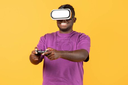 Foto de Retrato del hombre negro con gafas VR inmerso en la realidad aumentada utilizando el mando a distancia, disfrutar de juegos interactivos y explorar la realidad virtual sobre fondo de estudio amarillo - Imagen libre de derechos