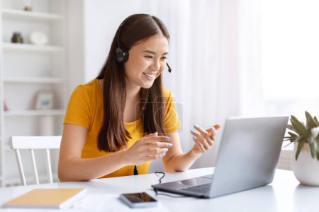 Photo for Portrait Of Smiling Asian Woman In Headset Making Video Call On Laptop, Happy Young Korean Female Sitting At Desk In Home Office And Talking At Computer Web Camera, Enjoying Teleconference - Royalty Free Image