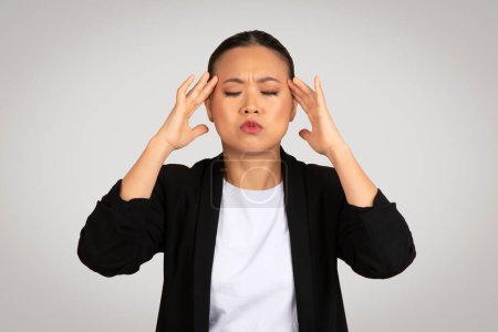 Photo for Stressed Asian businesswoman with a pained expression holding her head, depicting a headache or overwhelming pressure in a professional workplace scenario, isolated on gray background, studio - Royalty Free Image