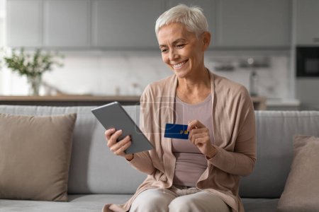 Photo for Easy Payment And Online Shopping. Happy mature lady holds digital tablet ordering purchases on internet, holding plastic credit card, seated on sofa in her modern living room - Royalty Free Image