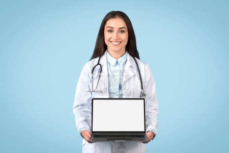 Téléchargez les photos : Heureux médecin européen femme montrant ordinateur portable avec écran vierge publicité offre médicale en ligne ou un service debout sur fond bleu studio, maquette - en image libre de droit