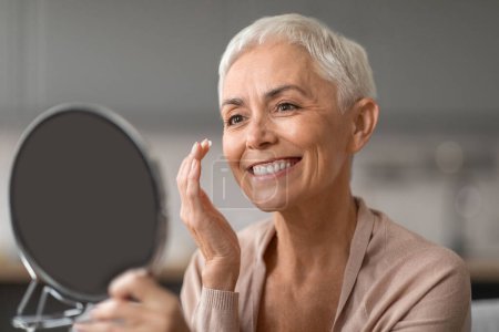 Photo for Anti Aging Skincare. Cheerful Senior Lady Applying Facial Moisturizer Preventing Wrinkles Looking At Round Mirror, Caring For Her Ageless Beauty At Home, Enjoying Self Pampering - Royalty Free Image