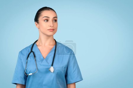 Foto de Retrato de la enfermera europea posando y mirando el espacio libre aislado sobre fondo de estudio azul, dama vistiendo abrigo azul y estetoscopio - Imagen libre de derechos