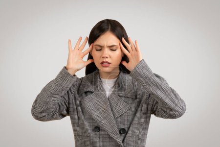 Téléchargez les photos : Jeune professionnelle européenne frustrée en manteau à carreaux ressentant des maux de tête, pressant ses doigts contre ses tempes dans un geste de stress ou de douleur - en image libre de droit