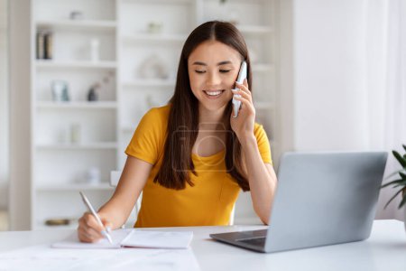 Photo for Home Office. Young Asian Woman Talking On Cellphone And Taking Notes While Working At Desk With Laptop Computer, Smiling Korean Freelancer Lady Enjoying Remote Job Opportunities, Free Space - Royalty Free Image
