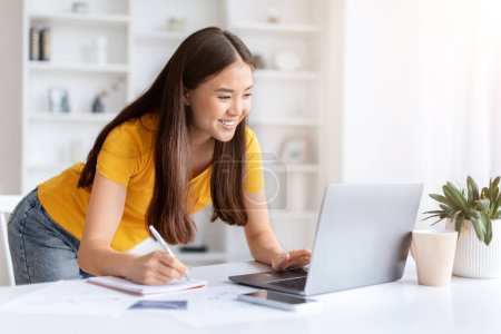 Foto de Mujer asiática enfocada trabajando en el ordenador portátil y tomando notas, sonriendo joven coreana apoyada en el escritorio en su oficina en casa, utilizando la computadora y anotando información para anotar, copiar espacio - Imagen libre de derechos