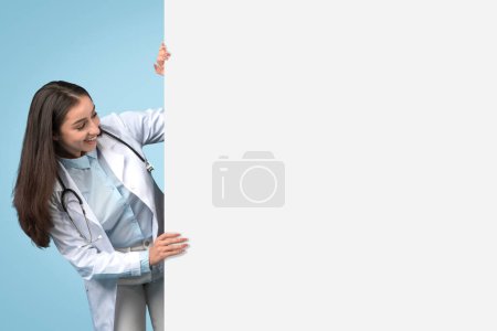 Foto de Doctora joven asomándose por detrás de una cartulina blanca en blanco para texto o diseño médico, doctora vestida con bata blanca con estetoscopio, espacio libre, pancarta - Imagen libre de derechos