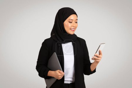 Photo for A modern professional Asian businesswoman in a hijab looks at her smartphone while holding a laptop, representing multitasking and technological proficiency, isolated on gray background, studio - Royalty Free Image