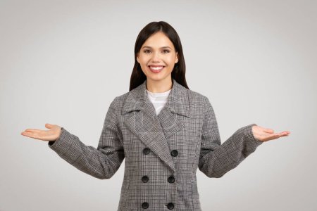 Photo for Joyful young woman in plaid blazer with hands outstretched, palms up, as if happily balancing or weighing different options against neutral grey backdrop - Royalty Free Image