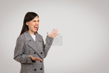 Furious young businesswoman in gray plaid blazer shouting and gesturing with her hand, expressing strong dissatisfaction or argument, free space