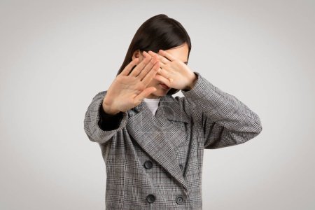 Photo for Stylish young woman in a houndstooth coat makes a see no evil gesture, covering her face with hands, symbolizing avoidance or privacy - Royalty Free Image
