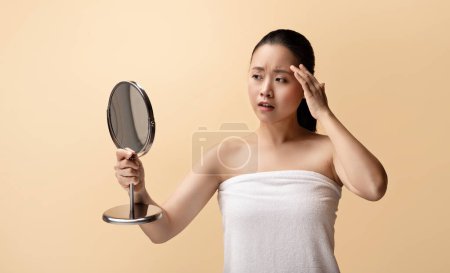 Téléchargez les photos : Triste dame nue chinoise millénaire en serviette, regarder miroir, a la peau problème, rides, isolé sur fond beige, studio. Soins de beauté naturels, hydratation de routine à la maison - en image libre de droit