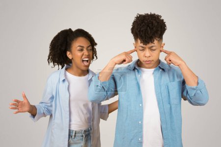 Zwei junge afrikanisch-amerikanische Individuen, eine fröhliche Dame, die lebhaft schreit, und ein Mann mit frustriertem Gesichtsausdruck, der seine Ohren mit den Fingern vor hellem Hintergrund blockiert