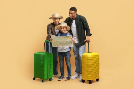 Photo for Family tourism. Father, mother and son checking paper map, looking for destination to travel, carrying suitcases, wearing wicker hats, isolated on beige studio background - Royalty Free Image