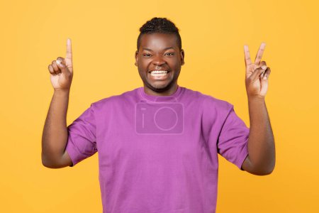 Foto de Feliz chico afroamericano levantando ambas manos mostrando uno y dos dedos, contando hasta tres con gestos, sonriendo a la cámara mientras está de pie sobre el fondo amarillo del estudio, retrato - Imagen libre de derechos