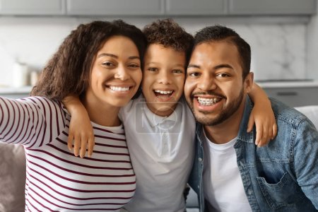 Foto de Feliz joven negro padres tomando selfie con pequeño hijo en casa, alegre afro-americano familia de tres con preadolescente macho niño captura foto juntos, mirando y sonriendo en cámara, primer plano - Imagen libre de derechos