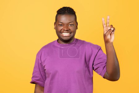Téléchargez les photos : Joyeux homme noir en t-shirt violet levant la main montrant deux doigts, geste de victoire ou signe de paix, sourire à la caméra tout en posant debout sur fond de studio jaune, prise de vue portrait - en image libre de droit