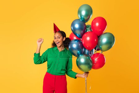 Foto de Fiesta de cumpleaños y sorpresas. Jovencita negra alegre sosteniendo un montón de globos brillantes, haciendo un gesto sí celebrando su día con alegría contra el fondo del estudio amarillo. Celebración festiva - Imagen libre de derechos