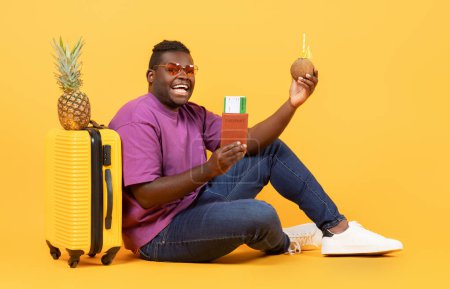 Téléchargez les photos : Heureux touriste afro-américain tenant des billets de voyage et une boisson à la noix de coco avec des pailles, assis près de sa valise avec de l'ananas dessus, prêt pour les vacances d'été, posant sur fond de studio - en image libre de droit