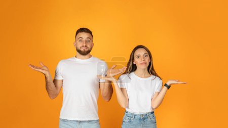 Photo for Confused young couple in white t-shirts and denim jeans shrugging with hands outstretched against a bright orange background, expressing uncertainty or indecision, panorama - Royalty Free Image
