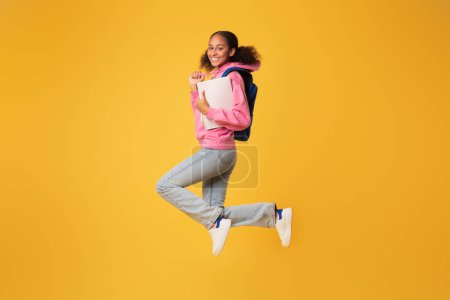 Foto de Sonriente niña afroamericana joven saltando con su mochila y libros de trabajo de la universidad, posando en el aire sobre el fondo del estudio amarillo. Largura completa tiro de despreocupado feliz estudiante - Imagen libre de derechos